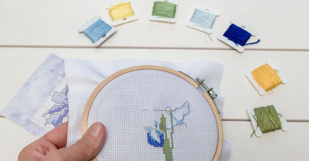 Blue and green embroidery floss skeins and hands cross stitching Aida in embroidery hoop